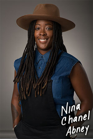 A headshot of woman with long dreadlocks, wearing a denim shirt and overalls. 