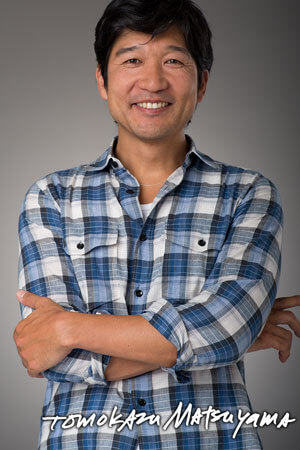 A portrait of a man smiling and wearing a blue, checkered shirt in front of a grey background. 