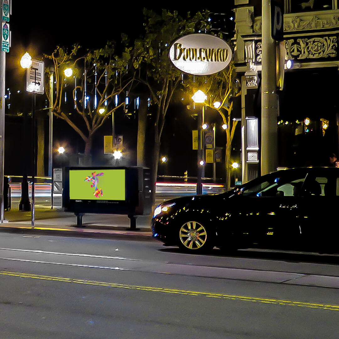 A black car on the street at night with a bright piece of artwork displayed on a box on the sidewalk.