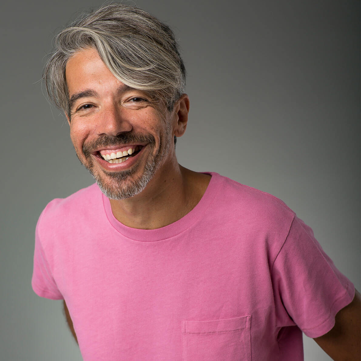 A headshot of a grey-haired man smiling and wearing a pink t-shirt.