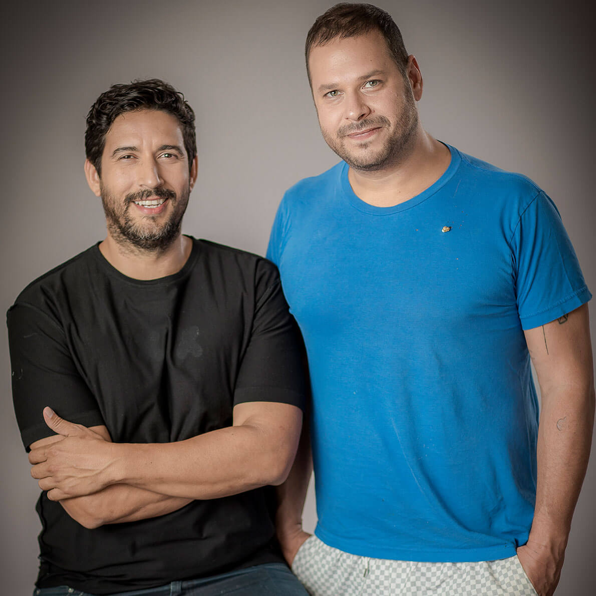 A studio portrait of two men. One of them is sitting on a chair and the other one is standing.