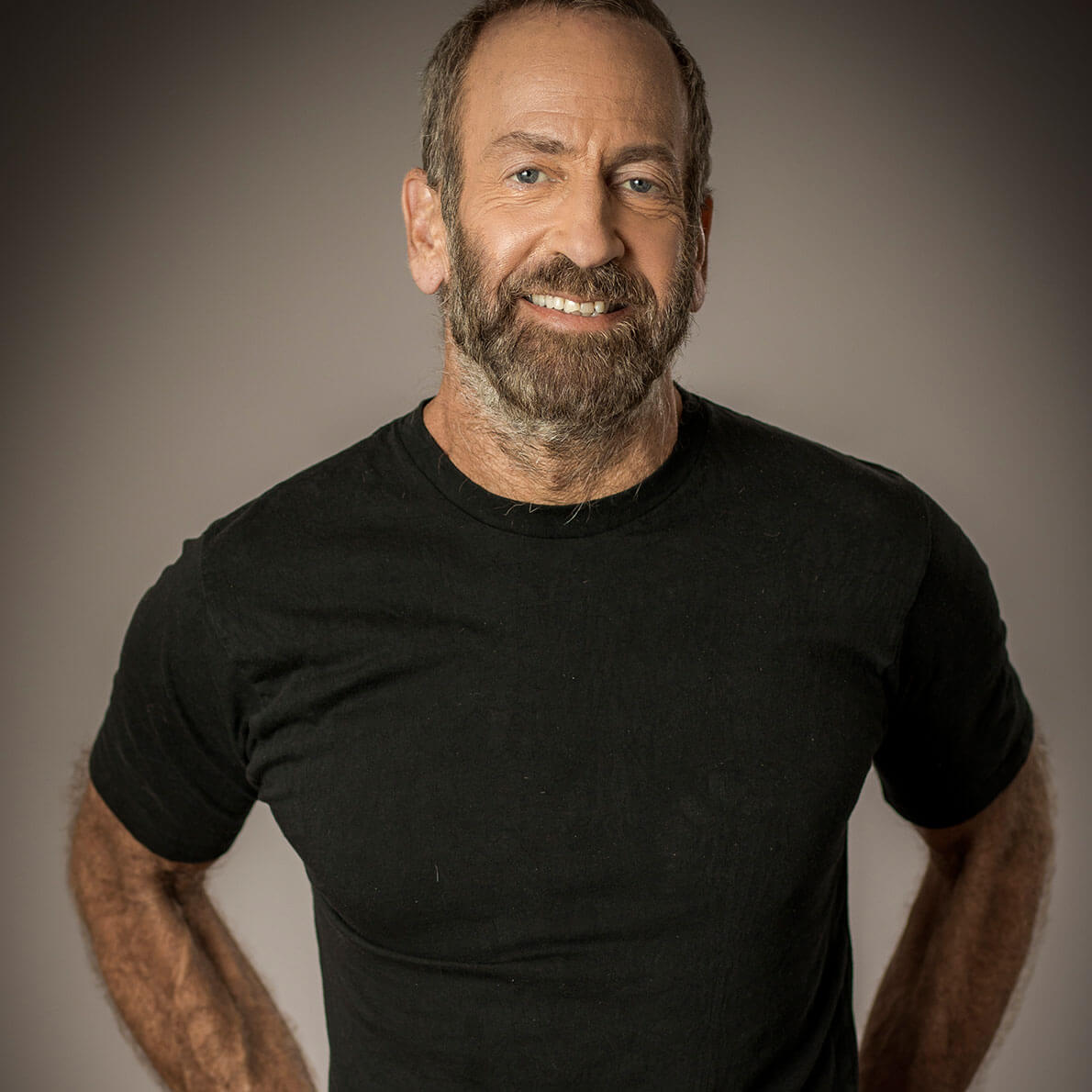 A portrait of a middle-aged man with a beard, wearing a black t-shirt and standing with his hands in his back pocket in front of a grey background.