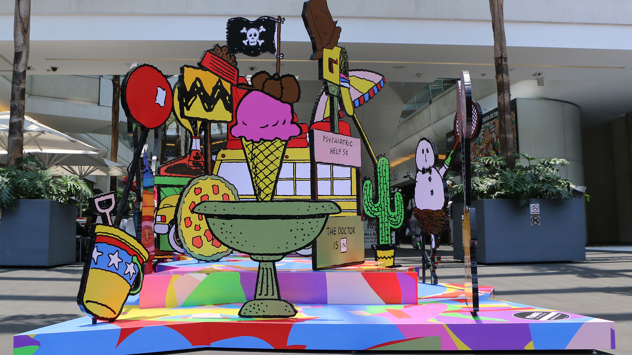 A colourful, 3D piece of art with many random objects including an icecream cone, a beach umbrella, and a school bus.