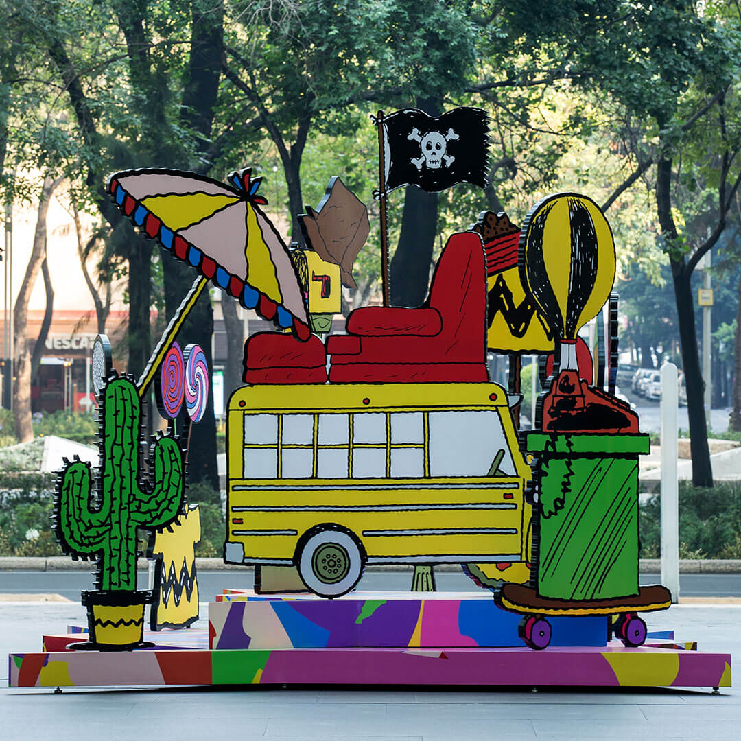 A colourful, 3D piece of art of a yellow school bus surrounded by many random items including a cactus, a couch and a beach umbrella. Behind the art installation is a park.