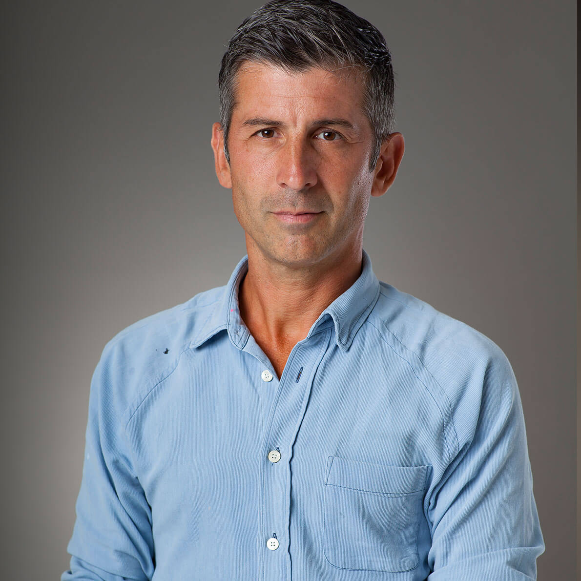 A portrait of a middle-aged man wearing a denim shirt in front of a grey background.