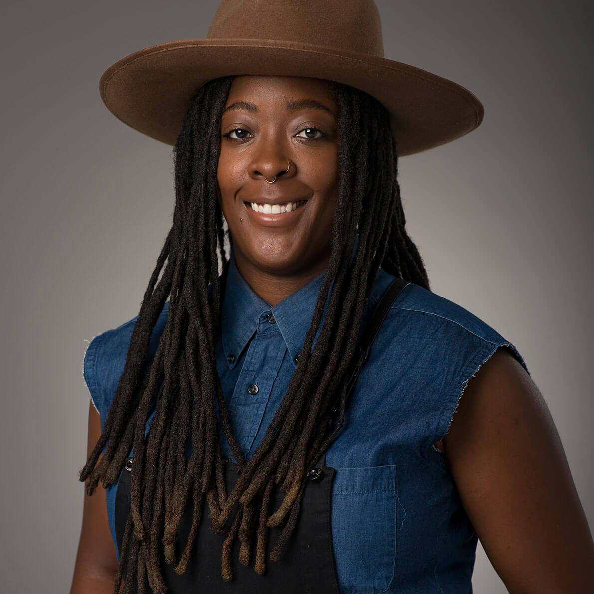 A headshot of woman with long dreadlocks, wearing a denim shirt and overalls.