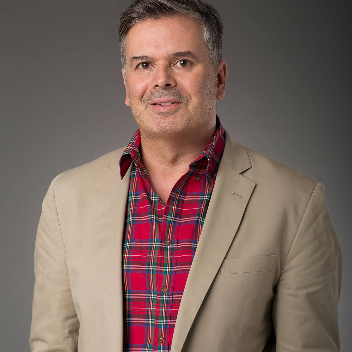 A portrait of a middle-aged man wearing a red shirt, brown blazer in front of a grey background.