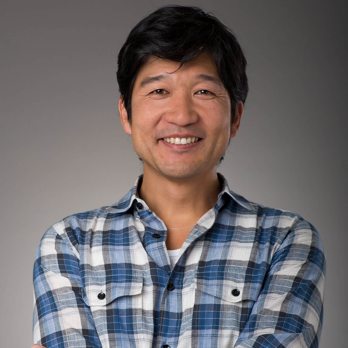 A portrait of a man smiling and wearing a blue, checkered shirt in front of a grey background.