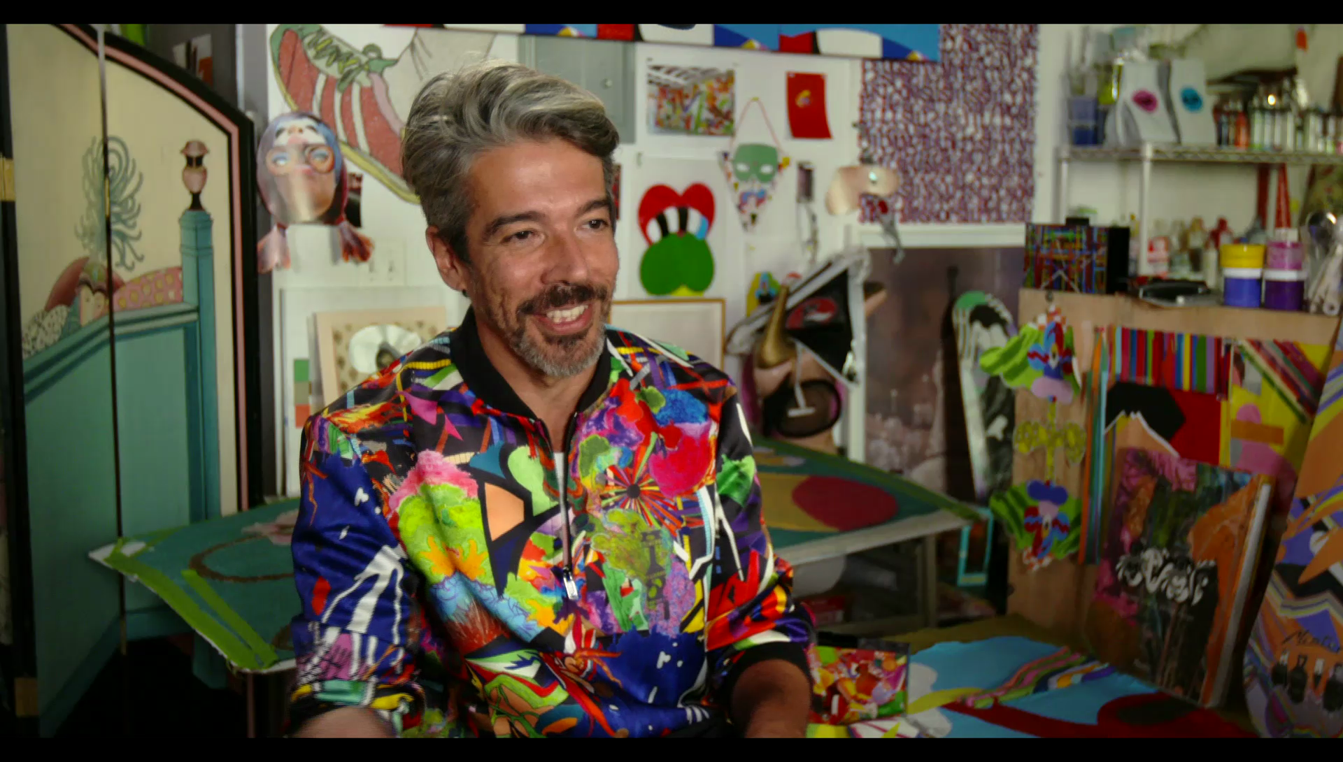 A man sitting in an art studio surrounded by paintings, wearing a colourful t-shirt.