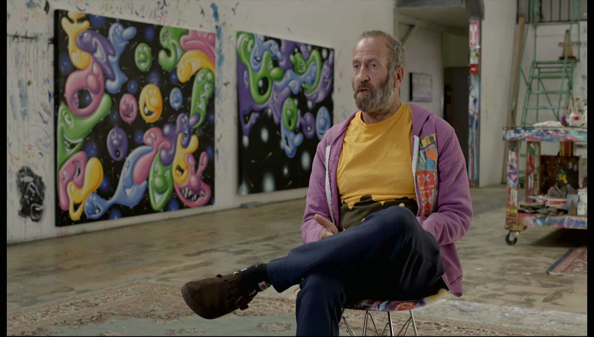 A man sitting in a chair, being interviewed in an art gallery.