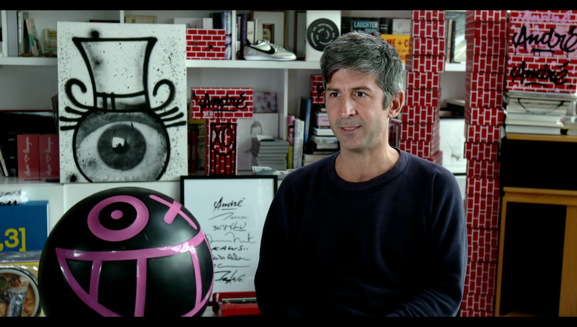 A man sitting in a room with artwork in the background, ready to be interviewed. 
