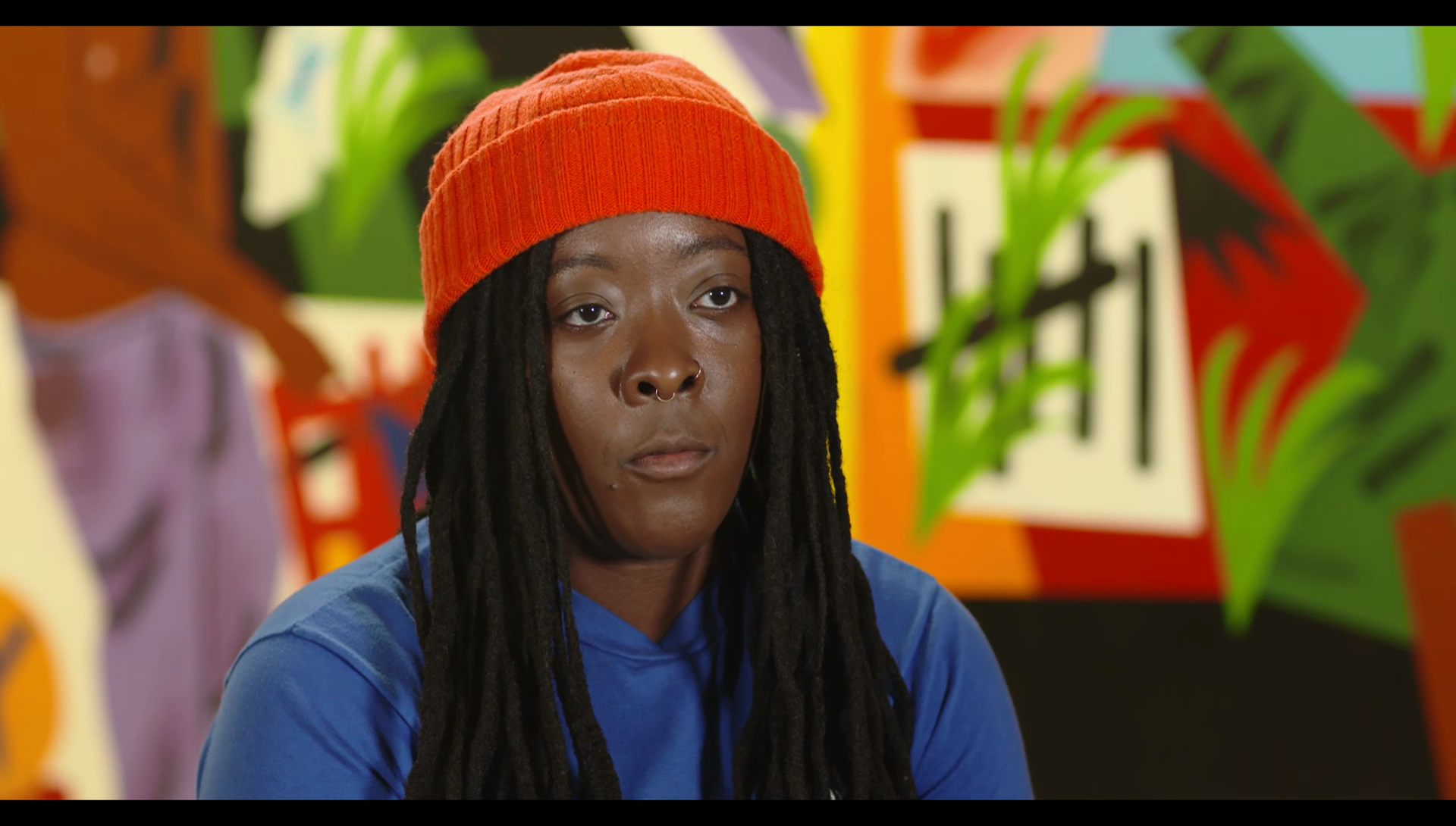 A close-up of a woman wearing a red hat, blue shirt, sitting in front of a colourful wall.