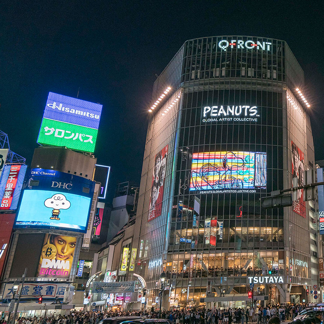 A busy city square at night, with bright lights and large LED screens displaying various images.