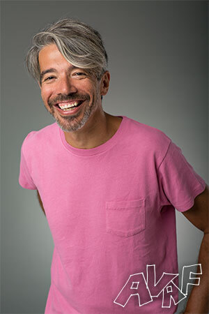A portrait of a grey-haired man, wearing a pink shirt in front of a grey background. 