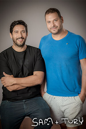 A studio portrait of two men. One of them is sitting on a chair and the other one is standing. 