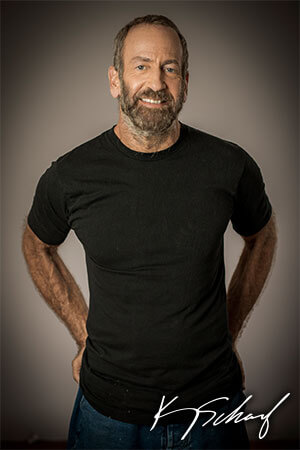 A portrait of a middle-aged man with a beard, wearing a black t-shirt and standing with his hands in his back pocket in front of a grey background. 