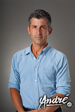 A portrait of a middle-aged man wearing a denim shirt in front of a grey background. 