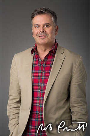 A portrait of a middle-aged man wearing a red shirt, brown blazer in front of a grey background. 