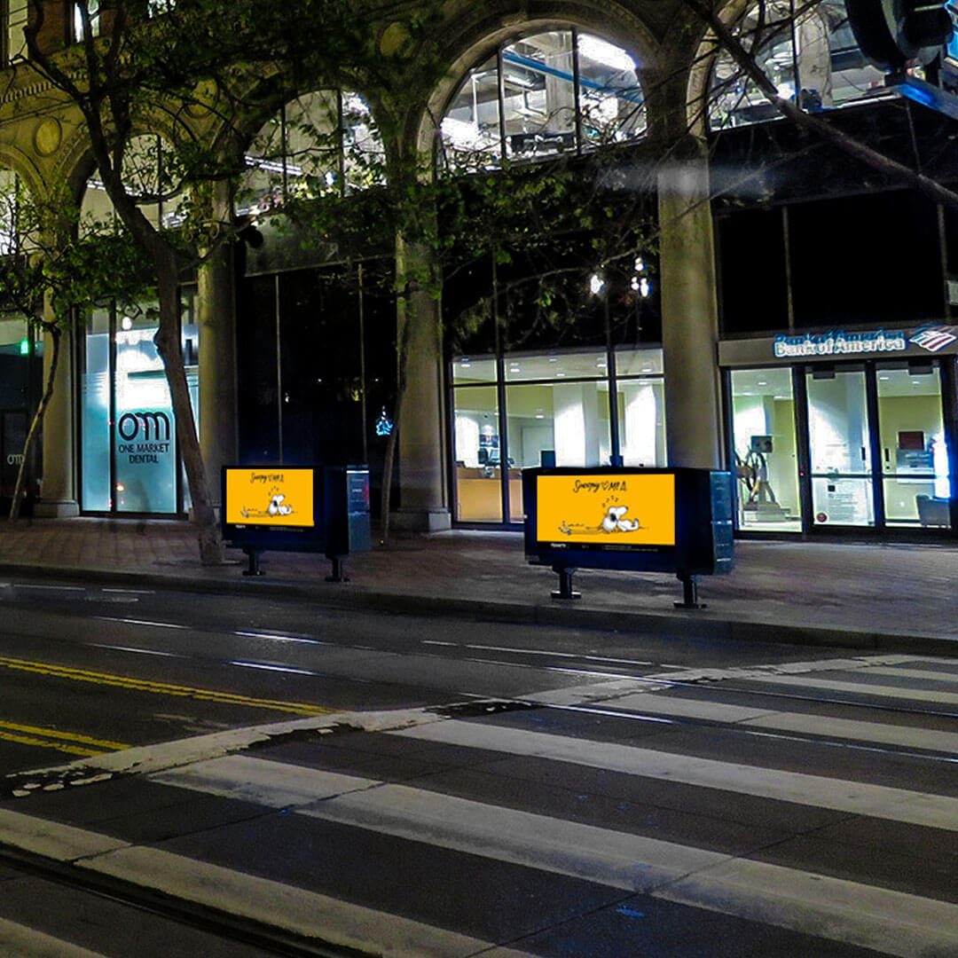 A street crossing at night with two boxes displaying bright pieces of art on the sidewalk.