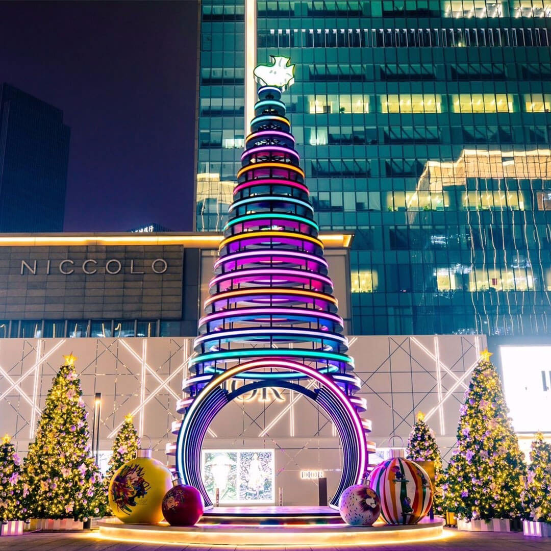 A brightly lit cone structure in front of a tall building, surrounded by Christmas trees.