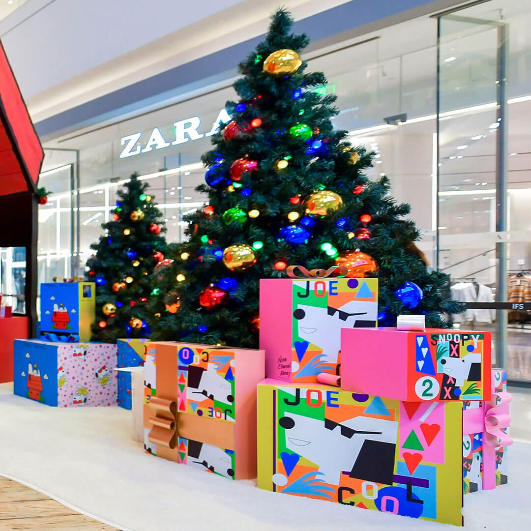 A bunch of Christmas presents sitting underneath two Christmas trees inside a shopping mall.