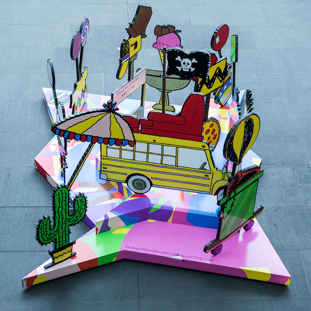 A colourful, 3D piece of art of a yellow school bus surrounded by many random items including a cactus, a couch and a beach umbrella. 