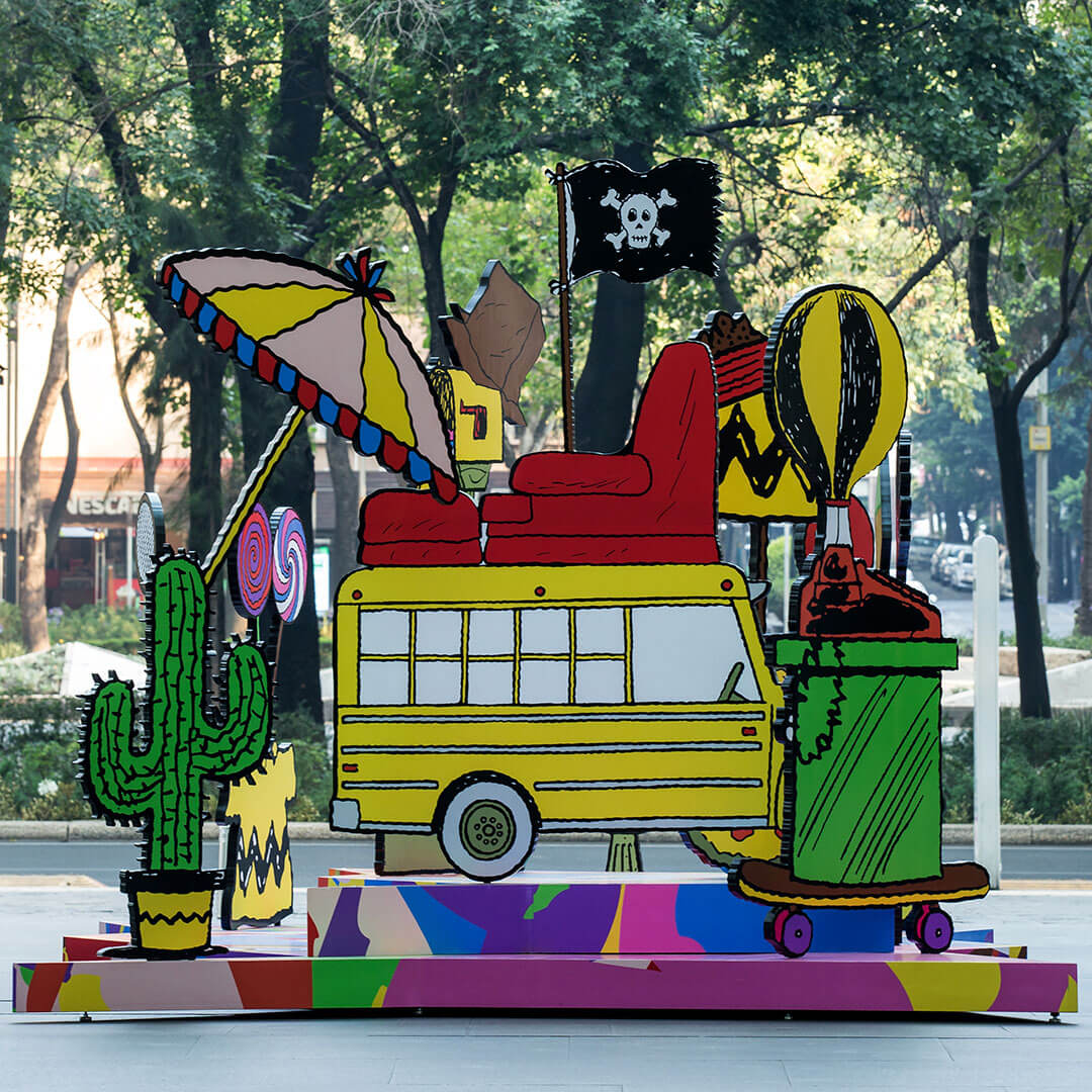 A colourful, 3D piece of art of a yellow school bus surrounded by many random items including a cactus, a couch and a beach umbrella. Behind the art installation is a park. 